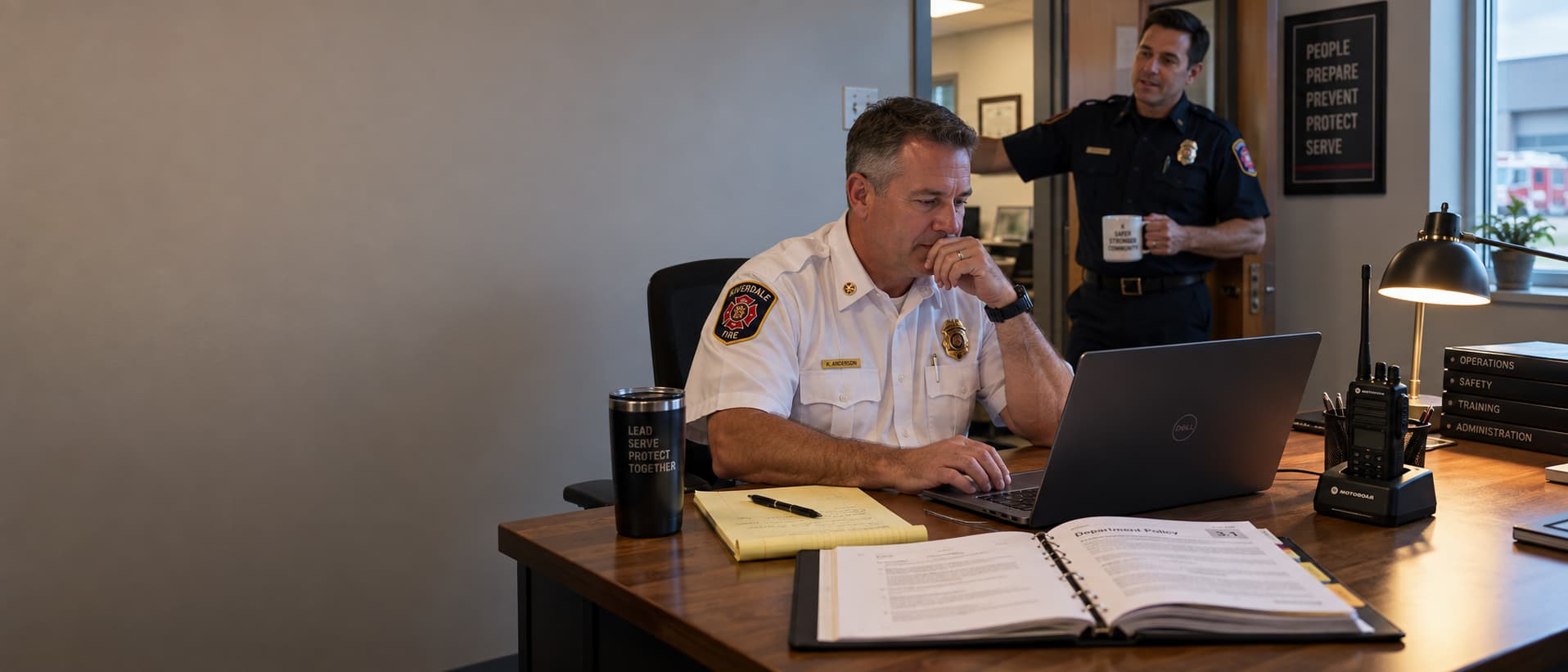 A fire chief at a station office desk completing a questionnaire on a laptop, with a legal pad and an open policy binder beside it.