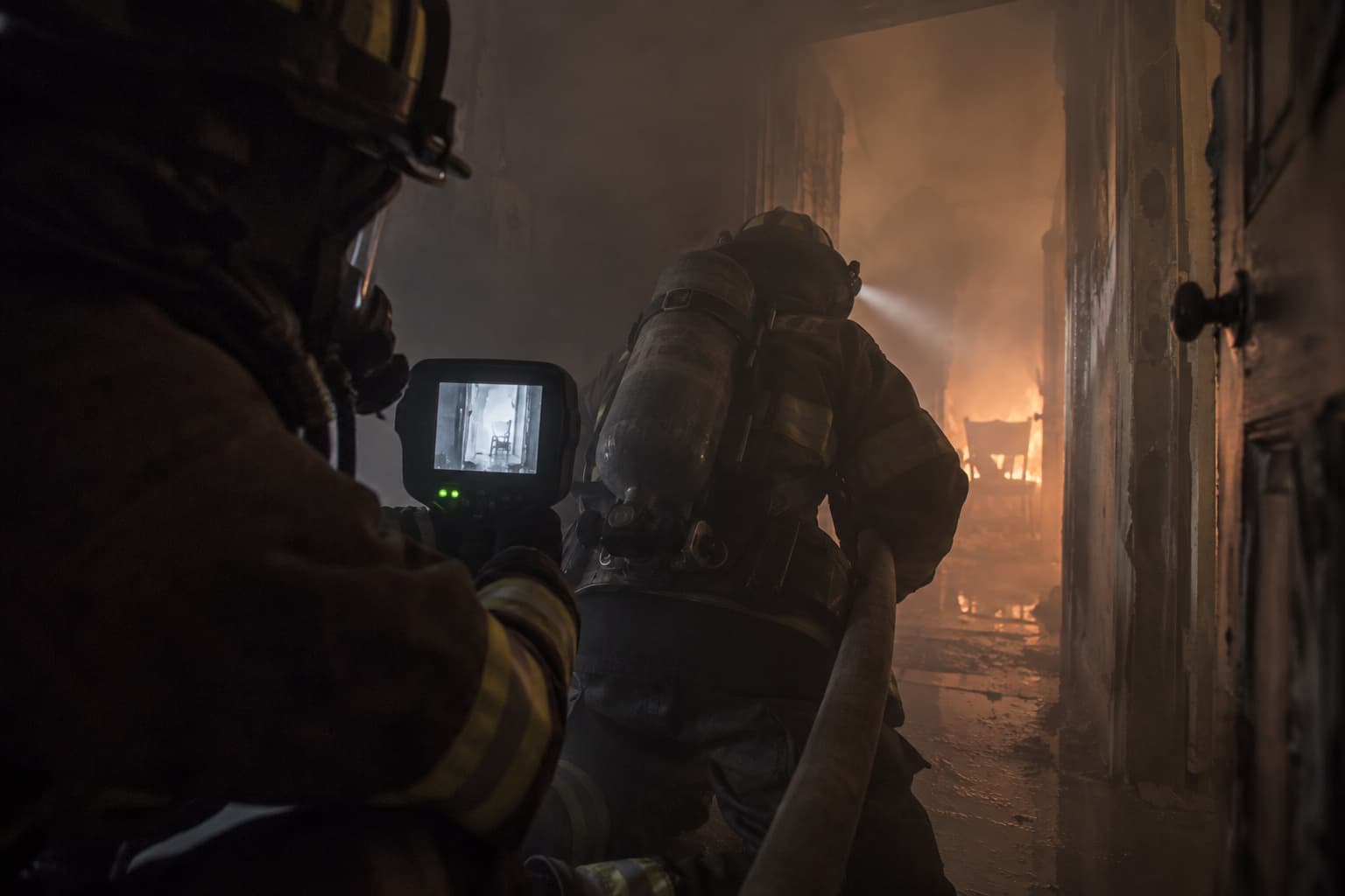 Interior view of a firefighter advancing a hoseline in heavy smoke, seen alongside the thermal image the crew is working from.